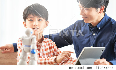 A boy moves a robot on a tablet and his father watches over him. 110433882