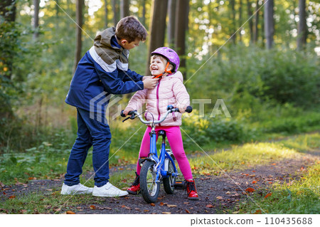 School kid boy, brother put on little preschool sister girl bike helmet on head. Brother teaching happy child cycling and having fun with learning bike. Active siblings family outdoors. Kids activity School kid boy, brother put on little preschool sister girl bike helmet on head. Brother teaching happy child cycling and having fun with learning bike. Active siblings family outdoors. Kids activity 110435688