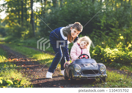 Two happy children playing with big old toy car in autumn forest, outdoors. Kid boy pushing and driving car with little toddler girl, cute sister inside. Laughing and smiling kids. Lovely family 110435702
