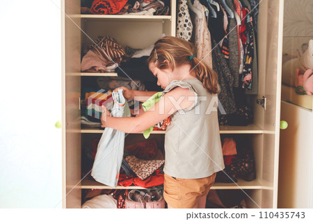 Little school girl standing by wardrobe with clothes. Preschool child putting shirts away and making decision for shirt to wear. Children get dressed in the morning for school. Little school girl standing by wardrobe with clothes. Preschool child putting shirts away and making decision for shirt to wear. Children get dressed in the morning for school. 110435743