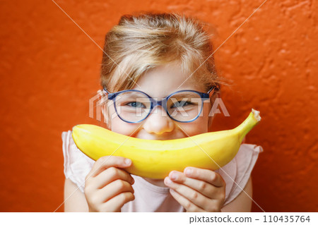 Happy little child girl with yellow banana like smile on orange background. Preschool girl with glasses smiling. Healthy fruits for children 110435764