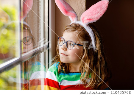 Portrait of a happy little girl with bunny ears looking outside sitting by a window. Easter holiday. Adorable child happy about holiday. Portrait of a happy little girl with bunny ears looking outside sitting by a window. Easter holiday. Adorable child happy about holiday. 110435768