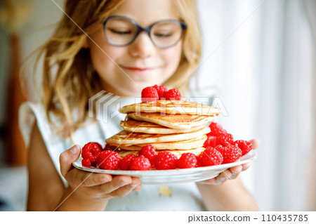 Little happy preschool girl with a large stack of pancakes and raspberries for breakfast. Positive child eating healthy homemade food in the morning. 110435785