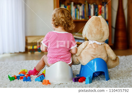 Closeup of cute little 12 months old toddler baby girl child sitting on potty. Kid playing with doll toy. Toilet training concept. Baby learning, development steps Closeup of cute little 12 months old toddler baby girl child sitting on potty. Kid playing with doll toy. Toilet training concept. Baby learning, development steps 110435792