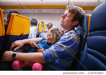 Young father holding his baby toddler daughter during flight on airplane going on vacations. Tired man and cute girl during night flight. child playing with smartphone. Family sitting in plane 110435863