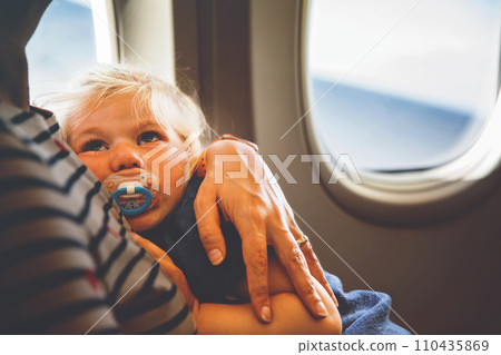 Woman traveling with little child by airplane. Sad tired toddler girl sitting with mum by aircraft window. Motherhood concept. Crying baby. 110435869