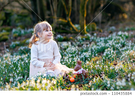 Little girl making Easter egg hunt in spring forest on sunny day, outdoors. Cute happy child with lots of snowdrop flowers and colored eggs. Springtime, christian holiday concept. 110435884