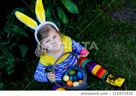 Cute little toddler girl with bunny ears having fun with traditional Easter eggs hunt on warm sunny day, outdoors. Happy child celebrating family christian holiday with basket with colored egg Cute little toddler girl with bunny ears having fun with traditional Easter eggs hunt on warm sunny day, outdoors. Happy child celebrating family christian holiday with basket with colored egg 110435916