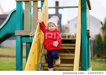 Cute toddler girl having fun on playground. Happy healthy little child climbing, swinging and sliding on different equipment. On cold day in colorful clothes. Active outdoors game for children Cute toddler girl having fun on playground. Happy healthy little child climbing, swinging and sliding on different equipment. On cold day in colorful clothes. Active outdoors game for children 110435933