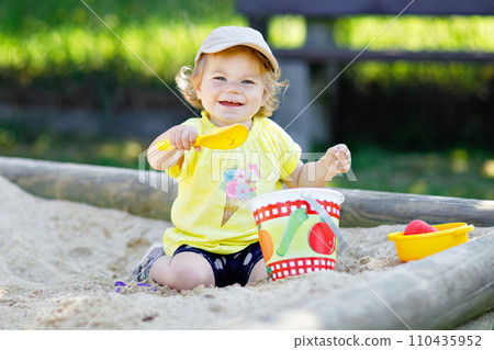 Cute toddler girl playing in sand on outdoor playground. Beautiful baby having fun on sunny warm summer sunny day. Happy healthy child with sand toys and in colorful fashion clothes. 110435952