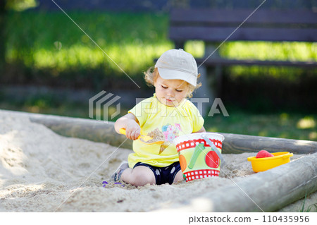 Cute toddler girl playing in sand on outdoor playground. Beautiful baby having fun on sunny warm summer sunny day. Happy healthy child with sand toys and in colorful fashion clothes. 110435956