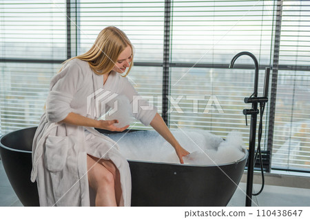 Spa relaxation. Young woman in spa bathrobe sitting at bath with foam in bathtub. Girl relaxing in bathroom at home. Female spending morning in bathroom. Beauty treatment body skin care concept Spa relaxation. Young woman in spa bathrobe sitting at bath with foam in bathtub. Girl relaxing in bathroom at home. Female spending morning in bathroom. Beauty treatment body skin care concept 110438647