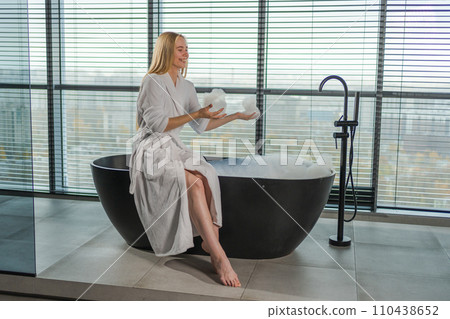Spa relaxation. Young woman in spa bathrobe sitting at bath with foam in bathtub. Girl relaxing in bathroom at home. Female spending morning in bathroom. Beauty treatment body skin care concept Spa relaxation. Young woman in spa bathrobe sitting at bath with foam in bathtub. Girl relaxing in bathroom at home. Female spending morning in bathroom. Beauty treatment body skin care concept 110438652