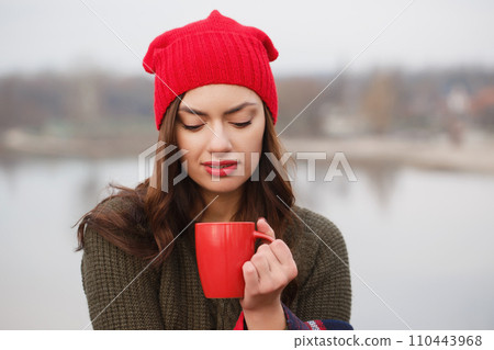 A young woman in a red hat and blanket with a red cup of hot drink 110443968