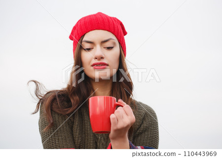 A young woman in a red hat and blanket with a red cup of hot drink 110443969