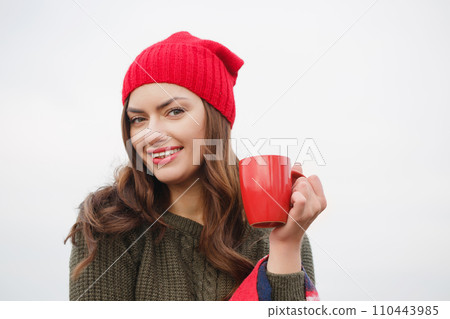 A young woman in a red hat and blanket with a red cup of hot drink 110443985