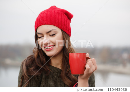 A young woman in a red hat and blanket with a red cup of hot drink 110443986