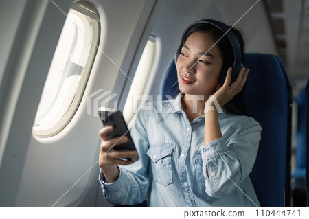 Asian woman traveler in airplane wearing headset listening music from mobile phone going on a trip vacation travel concept 110444741