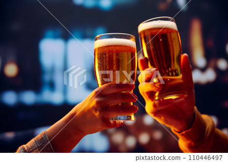 Hands holding lager light foamy big beer glasses and clinking. Two friends sitting in bar and drinking alcohol drinks. Hands holding lager light foamy big beer glasses and clinking. Two friends sitting in bar and drinking alcohol drinks. 110446497