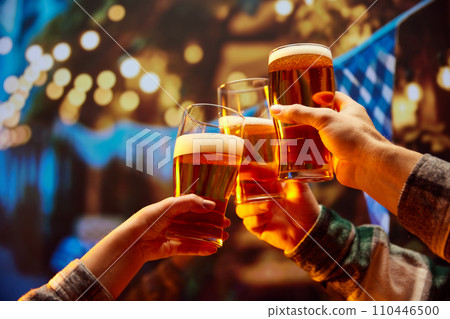 Close up image of hands holding glasses of beer and clinking after celebrating toast. Old friends sitting in bar and celebrating start of football season. Close up image of hands holding glasses of beer and clinking after celebrating toast. Old friends sitting in bar and celebrating start of football season. 110446500
