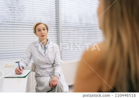 View from back to female doctor in white coat listening of unrecognizable woman patient tell about chronic disease, health complaints, affliction symptoms. Therapist take notes, fill history form. 110446518