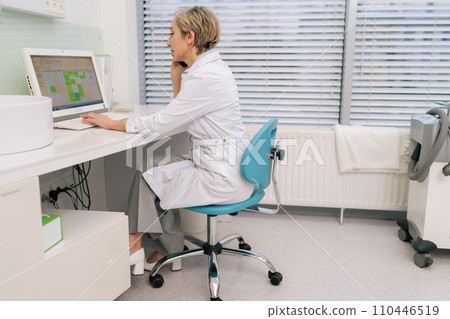Rear view of middle-aged woman general practitioner sit at desk in clinic office looking at laptop screen, discuss medical checkup results, talk to patient on cellphone work distantly use modern tech. 110446519
