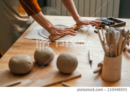 Artisan woman in lighted pottery studio, rolling out clay for modelling. Pottery studio with a variety of ceramics and a focused artisan Artisan woman in lighted pottery studio, rolling out clay for modelling. Pottery studio with a variety of ceramics and a focused artisan 110446933