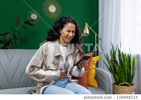 Sick unhealthy woman sitting on sofa at home, Hispanic woman having severe stomach pain, holding hands. 110448133