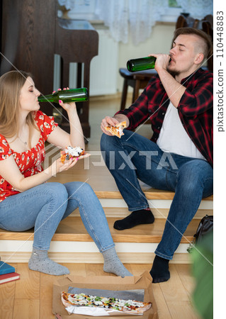 The couple is sitting on the floor with a pizza in a box in front of them. 110448883