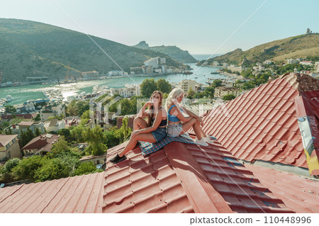Two women sitting on a red roof, enjoying the view of the town and the sea. Rooftop vantage point. In the background, there are several boats visible on the water, adding to the picturesque scene. 110448996