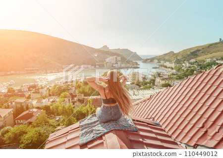 Woman sits on rooftop, enjoys town view and sea mountains. Peaceful rooftop relaxation. Below her, there is a town with several boats visible in the water. Rooftop vantage point. 110449012