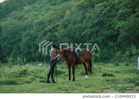 Happy blonde with horse in forest. Woman and a horse walking through the field during the day. Dressed in a plaid shirt and black leggings. 110449033