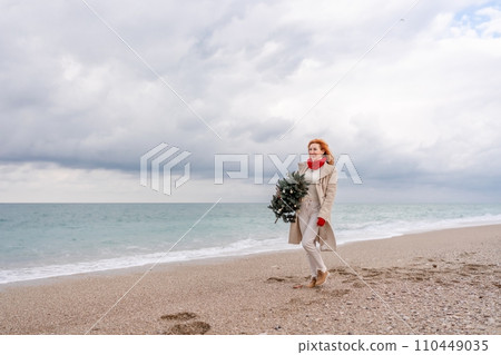 Redhead woman Christmas tree sea. Christmas portrait of a happy redhead woman walking along the beach and holding a Christmas tree in her hands. Dressed in a light coat, white suit and red mittens. 110449035