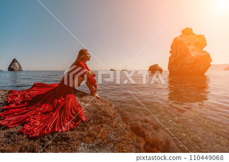 woman sea red dress. Beautiful sensual woman in a flying red dress and long hair, sitting on a rock above the beautiful sea in a large bay. 110449068