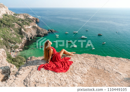 Woman sea red dress yachts. A beautiful woman in a red dress poses on a cliff overlooking the sea on a sunny day. Boats and yachts dot the background. 110449105
