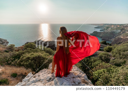 Woman sunset sea red dress, back view a happy beautiful sensual woman in a red long dress posing on a rock high above the sea on sunset. Woman sunset sea red dress, back view a happy beautiful sensual woman in a red long dress posing on a rock high above the sea on sunset. 110449179