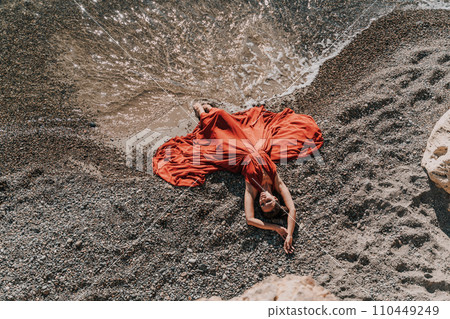 Woman red dress sea. Female dancer in a long red dress posing on a beach with rocks on sunny day. Girl on the nature on blue sky background. 110449249