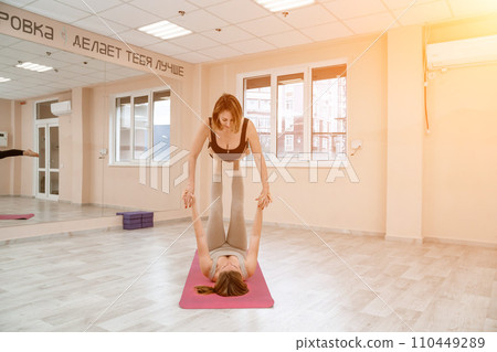 Two women do acroyoga on a light floor near a large mirror in a yoga studio. They are wearing leggings and tops. Two women do acroyoga on a light floor near a large mirror in a yoga studio. They are wearing leggings and tops. 110449289