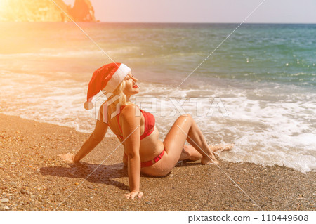 Female beach Santa hat wave coast. beach relaxation seaside. A woman in a red swimsuit enjoying her time on the beach, sits on the sand and being covered by a wave. Female beach Santa hat wave coast. beach relaxation seaside. A woman in a red swimsuit enjoying her time on the beach, sits on the sand and being covered by a wave. 110449608