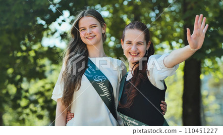 Two Russian schoolgirls graduate posing on a summer day with a ribbon and the name - Graduate 2023 on it. 110451227