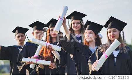 Cheerful graduates pose with raised diplomas on a sunny day. Cheerful graduates pose with raised diplomas on a sunny day. 110451233
