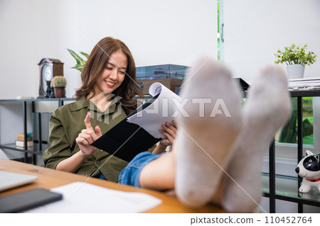 Working on financial. Businesswoman sitting relaxing reclining with feet up on computer desk reading report document in paperwork clipboard, smiling woman holding papers document reading with laptop 110452764