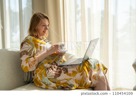 Pregnant woman, in a flowered dress, drinking tea and using a laptop sitting in the hotel hall. Pregnant woman, in a flowered dress, drinking tea and using a laptop sitting in the hotel hall. 110454109