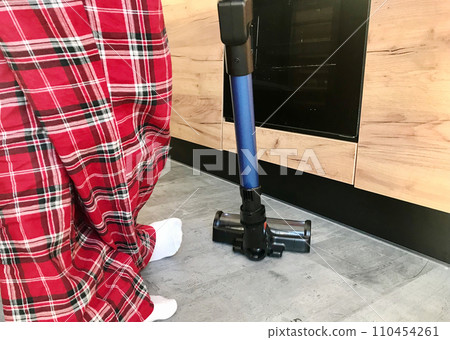 A woman in checkered pajamas is cleaning the gray floor with a vacuum cleaner 110454261