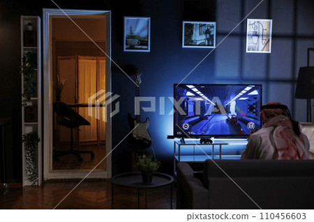 Rear-view shot of Muslim man seated on the sofa engrossed in playing video games. Image shows a Middle Eastern man dressed in traditional clothing enjoying online entertainment from the television. 110456603