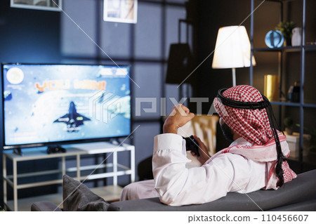 Youthful Arab man celebrates victory in virtual competition, controlling the action with a controller. The room is lit by the glow of the television as he enjoys an evening of digital entertainment. 110456607