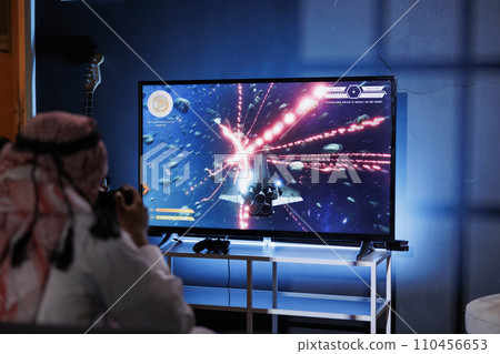 A Middle Eastern man playing a digital game at home. Immersed in cyberspace, he controls the game with a joystick, sitting in front of a detailed television screen. A Middle Eastern man playing a digital game at home. Immersed in cyberspace, he controls the game with a joystick, sitting in front of a detailed television screen. 110456653