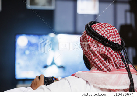 A young Muslim man celebrates victory while immersed in an online gaming competition at home. Selective focus of Arab guy grasping his wireless controller engaged in a video game. A young Muslim man celebrates victory while immersed in an online gaming competition at home. Selective focus of Arab guy grasping his wireless controller engaged in a video game. 110456674