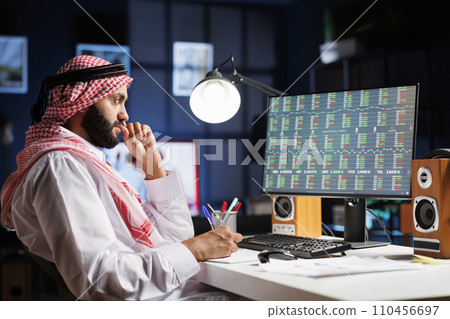 Diligent Muslim man working at a desk with a computer, checking financial reports and taking notes. Focused Arab guy using desktop pc, working with documents at the home office. 110456697
