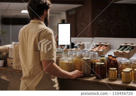 Client in zero waste shop using mockup tablet to videocall his girlfriend, buying her vegan food. Man with isolated screen device phone calls partner over internet in local supermarket 110456720
